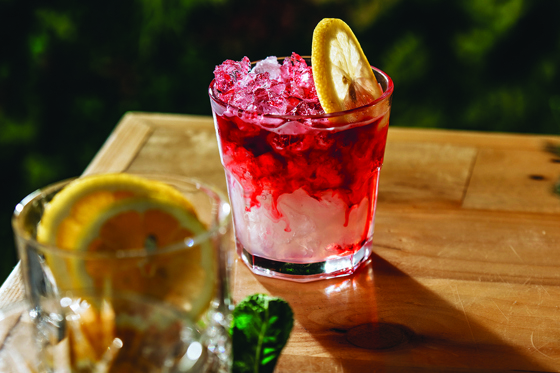 Cocktail with red colouring trickling down and a slice of lemon, with a glass full of lemons in foreground